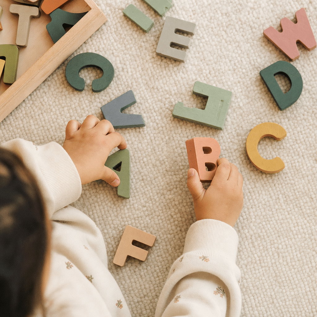 Mains d'un enfant manipulant des lettres en bois colorées sur un tapis clair pour apprendre l'alphabet.